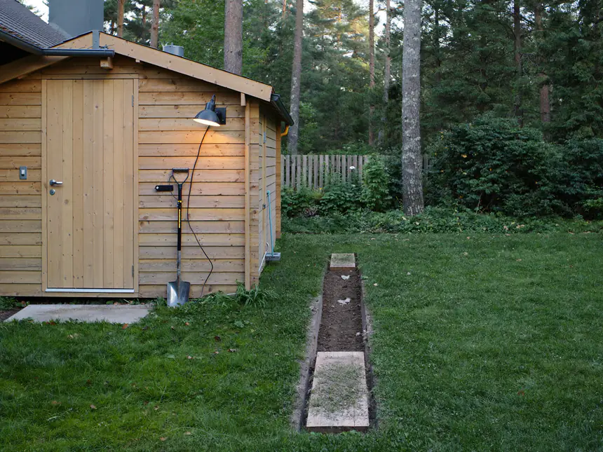 Houten tuinschuur aan de bosrand met een nieuwe buitenlamp en een sleuf voor de grondkabel door het gras