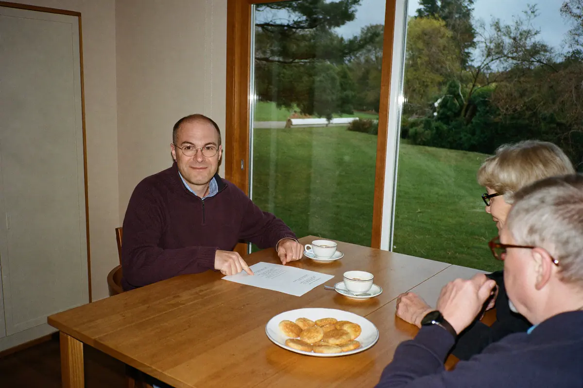Elektricien bespreekt aan de keukentafel het meetrapport met een ouder echtpaar, met uitzicht op een tuin met dennen